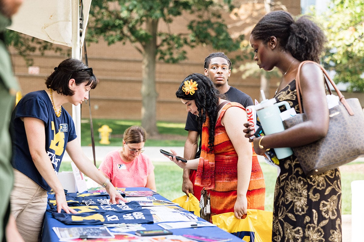 FLINT_M'Gagement Fair 2026_0022 A group of people interacting at a table with various items, including t-shirts and brochures, in an outdoor setting. One woman is examining items on the table, while another is looking at her phone. A man observes in the background.
