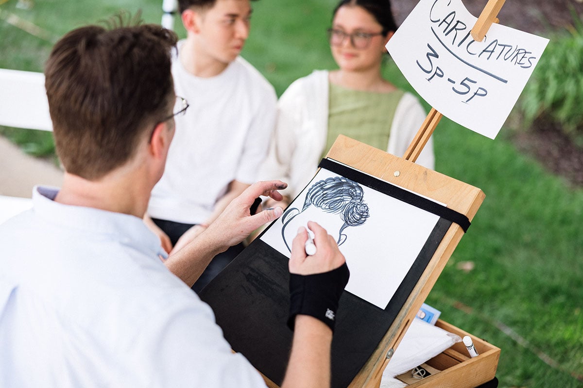 FLINT_M'Gagement Fair 2026_0026 A caricature artist draws a portrait on a sketchpad while two people sit nearby, observing. A sign above reads "CARICATURES 3P-5P."