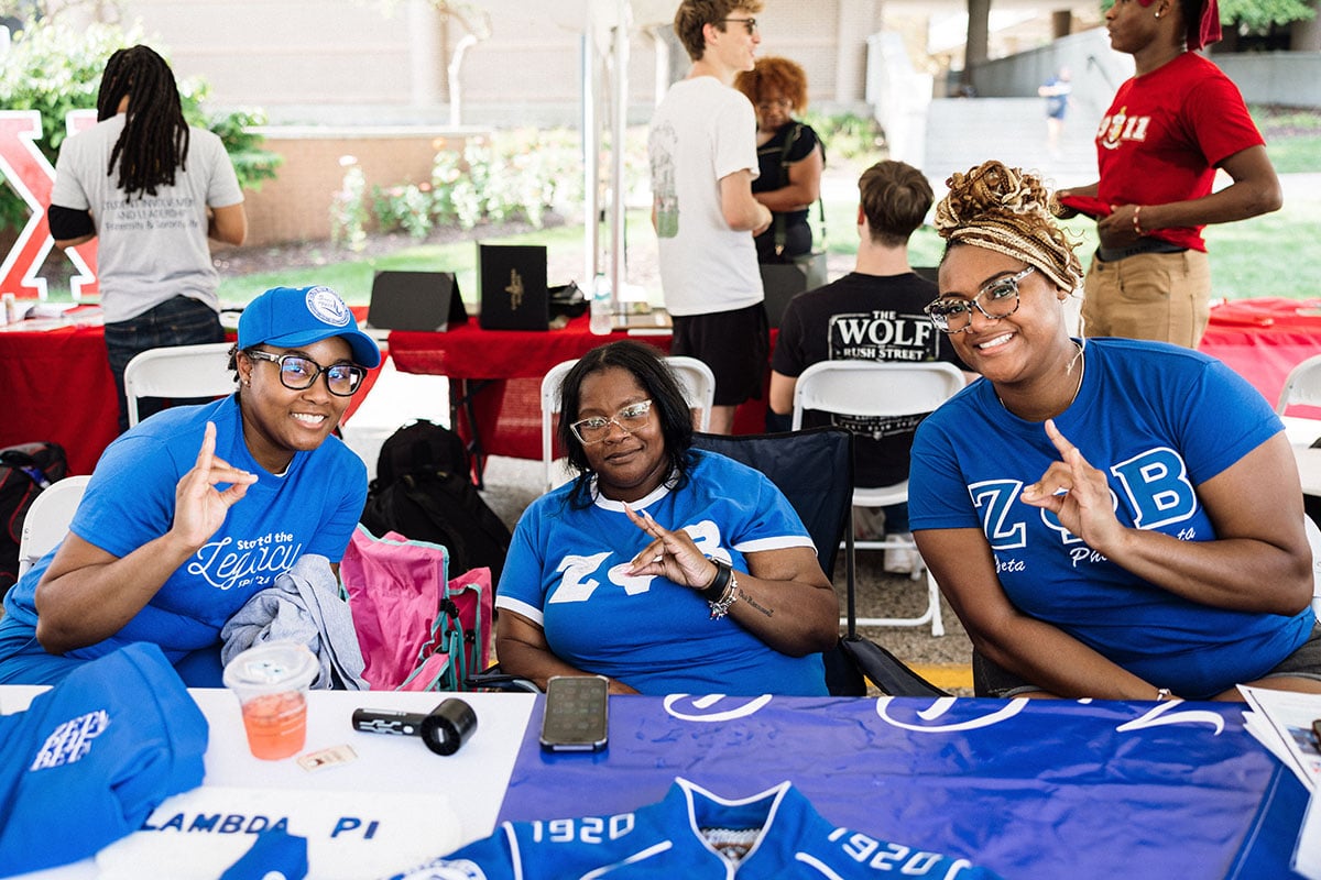 FLINT_M'Gagement Fair 2026_0029 Three women in blue shirts are smiling and making hand signs at a table with a banner and merchandise.