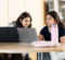 Two young women are sitting at a round table, focused on a laptop. One has a pink water bottle and a light pink laptop sleeve, while the other has a black laptop with a leopard print design.