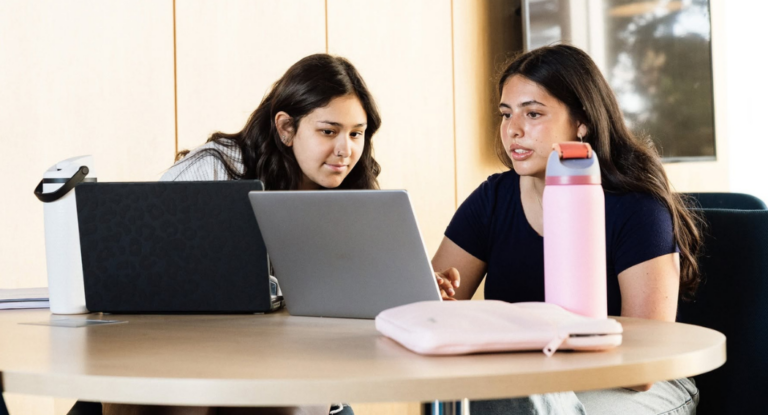 Two young women are sitting at a round table, focused on a laptop. One has a pink water bottle and a light pink laptop sleeve, while the other has a black laptop with a leopard print design.