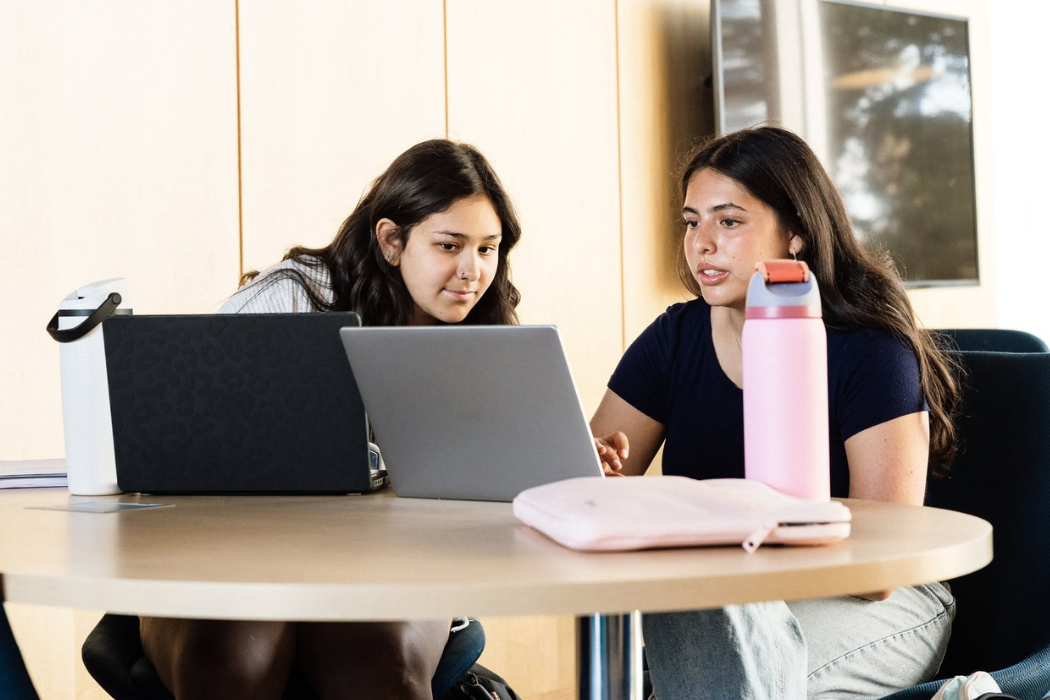 Two young women are sitting at a round table, focused on a laptop. One has a pink water bottle and a light pink laptop sleeve, while the other has a black laptop with a leopard print design.