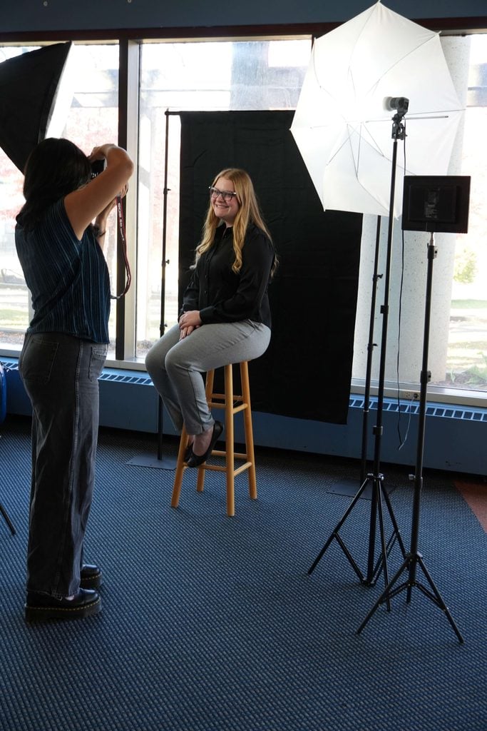 A photographer captures the portrait of a smiling young woman sitting on a stool in front of a black backdrop. Studio lighting is set up around her.