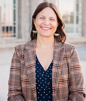 A woman with long brown hair smiles while wearing a patterned blazer and a polka dot dress, standing outdoors.