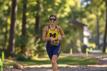 Sia Hayes, a female athlete wearing a blue and yellow racing suit with the letter "M" is running on a wooded path, smiling and looking ahead.