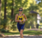 Sia Hayes, a female athlete wearing a blue and yellow racing suit with the letter "M" is running on a wooded path, smiling and looking ahead.