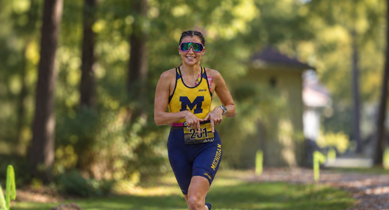 Sia Hayes, a female athlete wearing a blue and yellow racing suit with the letter "M" is running on a wooded path, smiling and looking ahead.