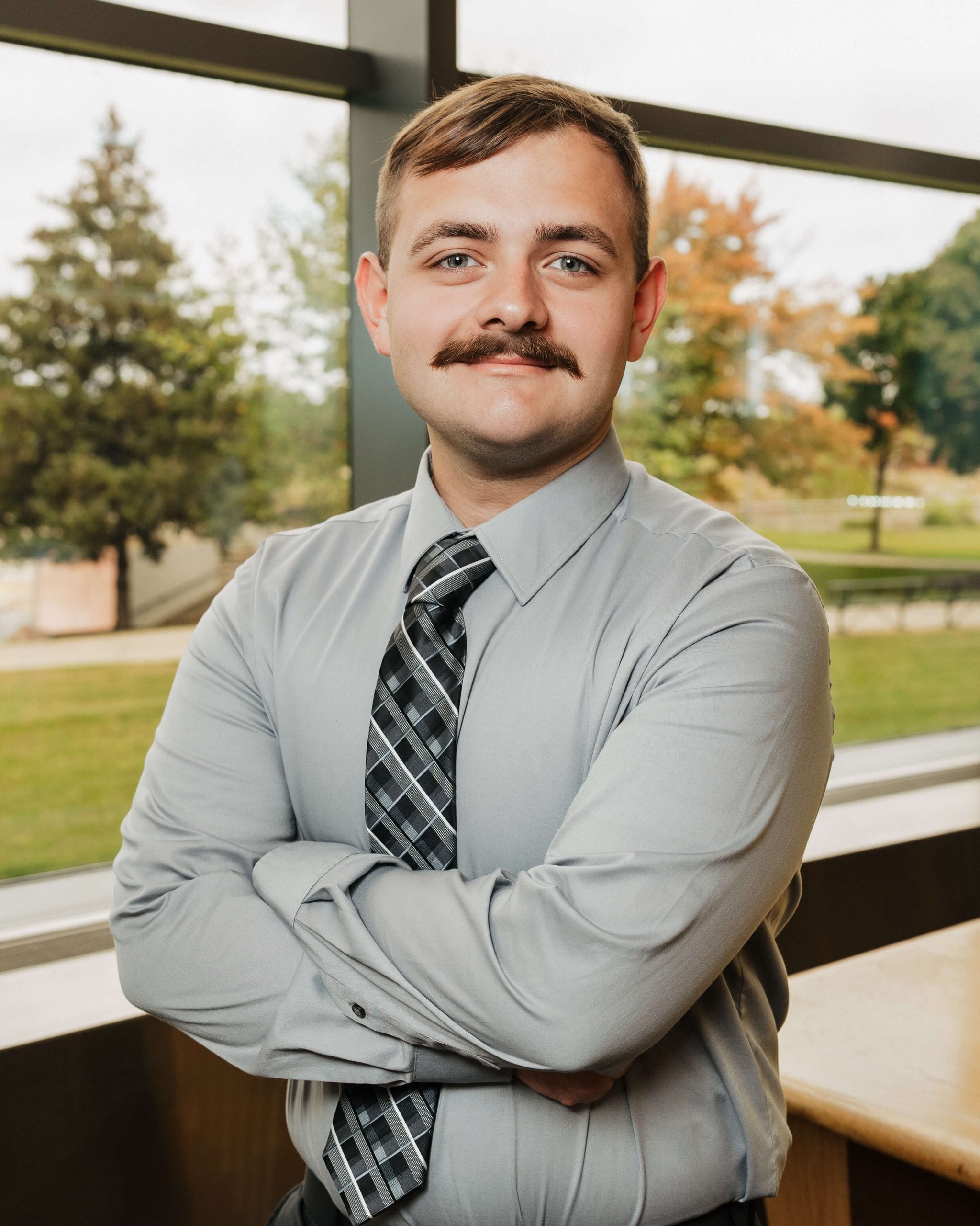 A professional portrait of a young man with a mustache wearing a button-up shirt and tie with his arms crossed. The background features tall windows through which one can see a park with trees.
