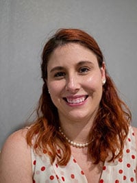 A woman with shoulder-length red hair, wearing a white dress with red polka dots and a pearl necklace, smiles at the camera.
