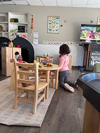 A child kneels at a small table with two dolls, surrounded by toys in a playroom. A television displays a colorful cartoon in the background.