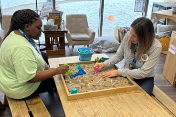 Two women are playing in a sand table, using colorful tools and toys. One woman is wearing a green shirt and the other a gray shirt with a "DIVERSITY" print.