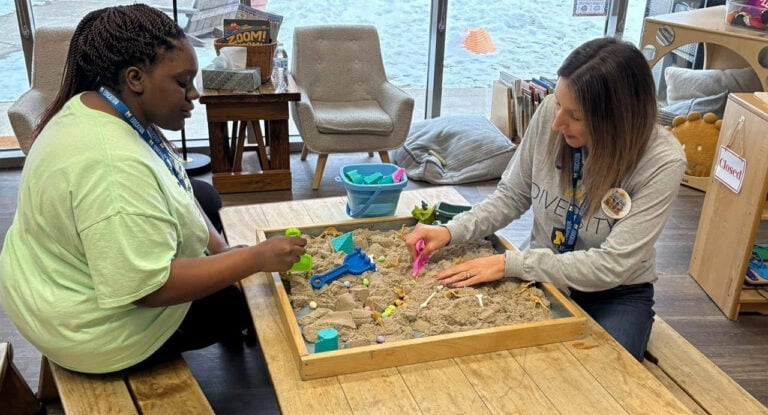 Two women are playing in a sand table, using colorful tools and toys. One woman is wearing a green shirt and the other a gray shirt with a "DIVERSITY" print.