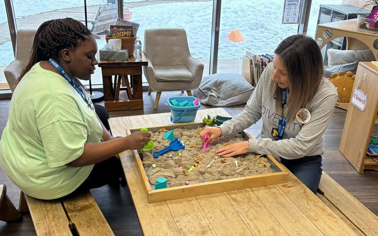 Two women are playing in a sand table, using colorful tools and toys. One woman is wearing a green shirt and the other a gray shirt with a "DIVERSITY" print.