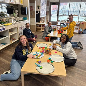 Five people are sitting on the floor around two tables in a classroom, engaged in activities with colorful play materials.