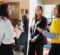 A group of three women are engaged in conversation at a career fair. One woman in a white sweater holds a flyer while another in a black blazer listens. A third woman in a yellow top gestures with her hands while speaking.