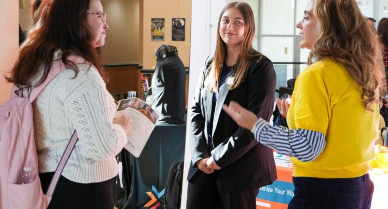 A group of three women are engaged in conversation at a career fair. One woman in a white sweater holds a flyer while another in a black blazer listens. A third woman in a yellow top gestures with her hands while speaking.