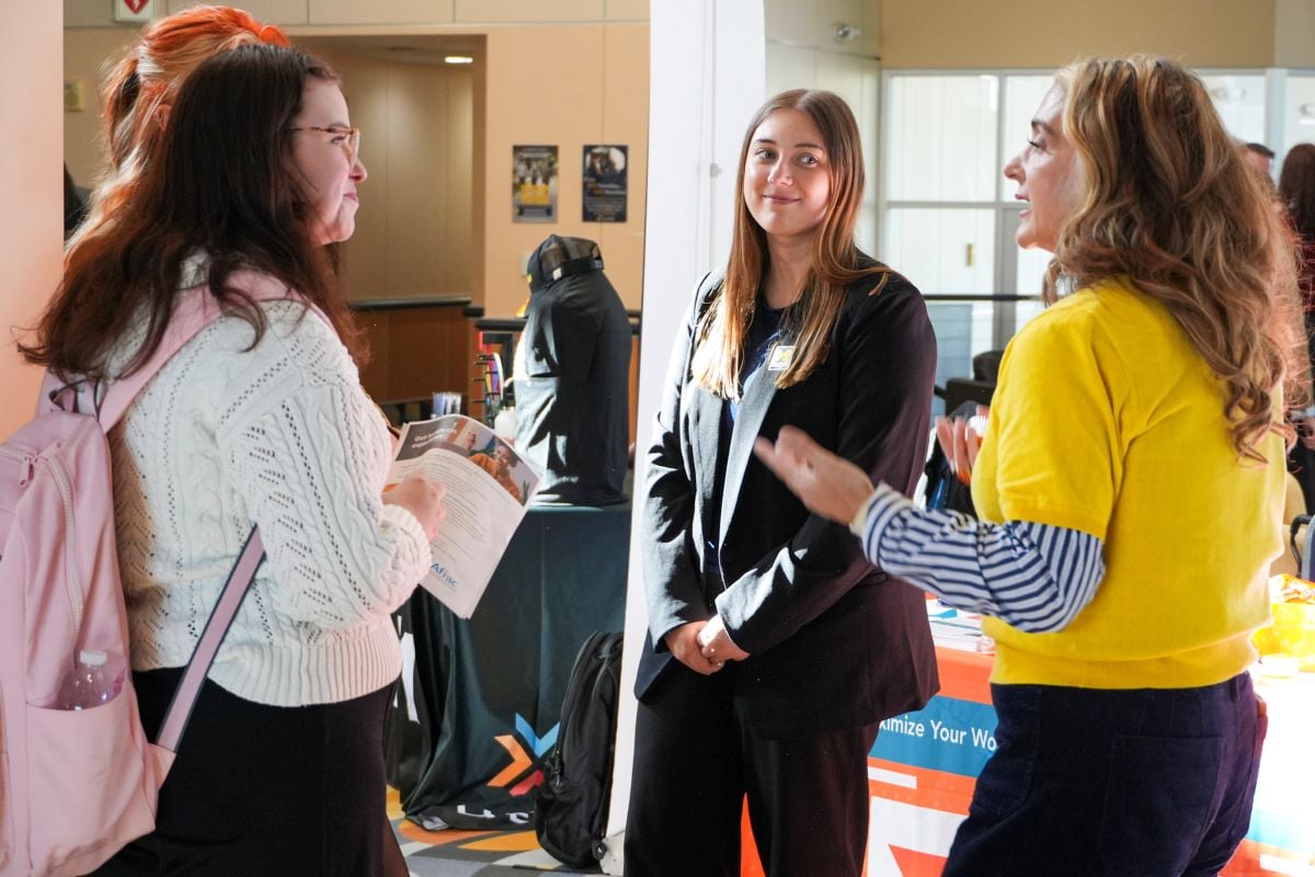 A group of three women are engaged in conversation at a career fair. One woman in a white sweater holds a flyer while another in a black blazer listens. A third woman in a yellow top gestures with her hands while speaking.