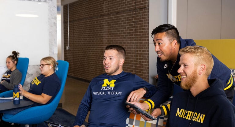 A group of students in a study area, three men engaged with a tablet, while three women sit nearby, all wearing university apparel.