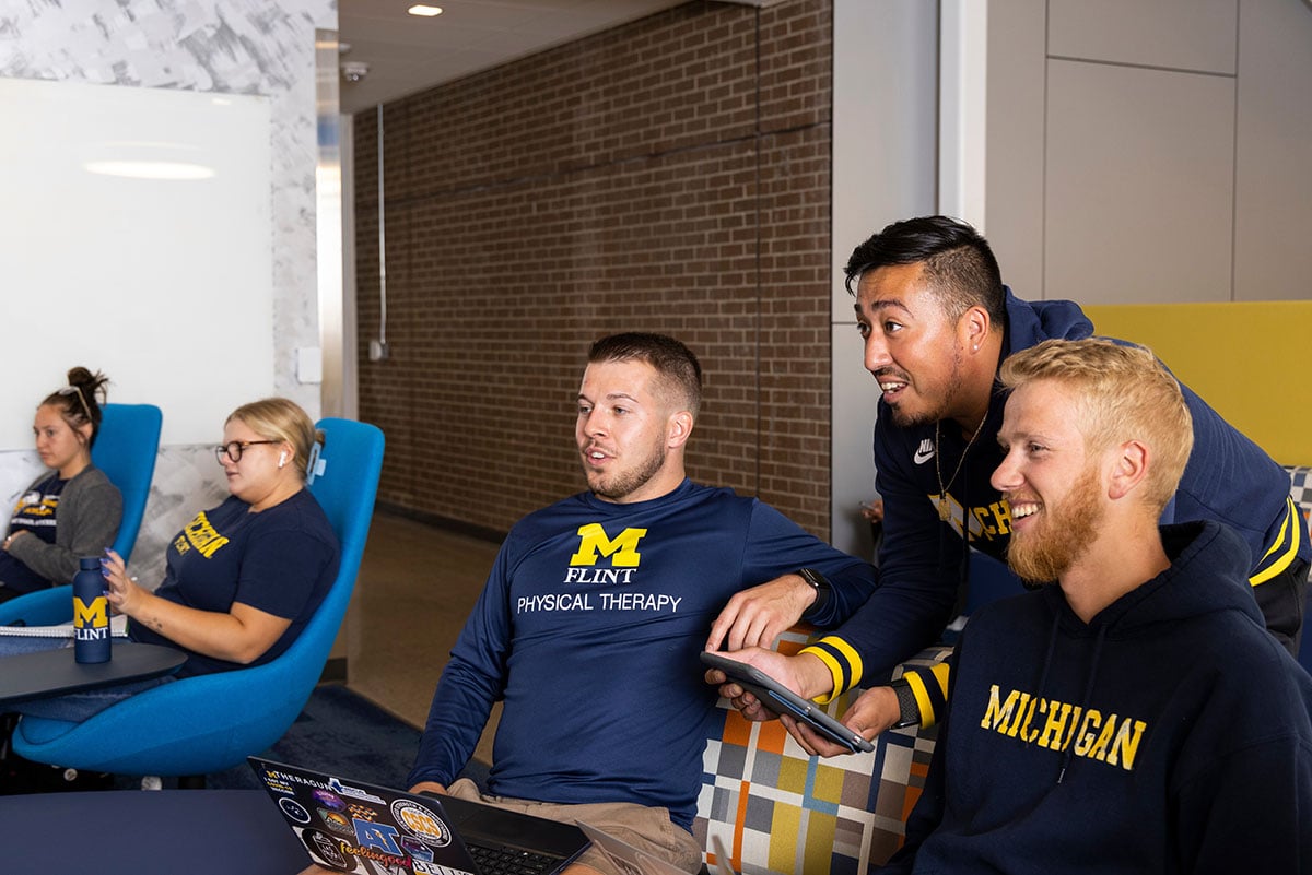 A group of students in a study area, three men engaged with a tablet, while three women sit nearby, all wearing university apparel.