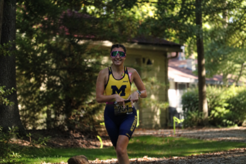 A smiling female athlete in a yellow and blue racing suit runs along a path surrounded by trees, wearing sunglasses and a race number.