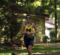 A smiling female athlete in a yellow and blue racing suit runs along a path surrounded by trees, wearing sunglasses and a race number.