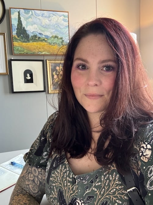 A woman with long, dark hair is smiling at the camera, sitting in front of a wall adorned with framed artwork.