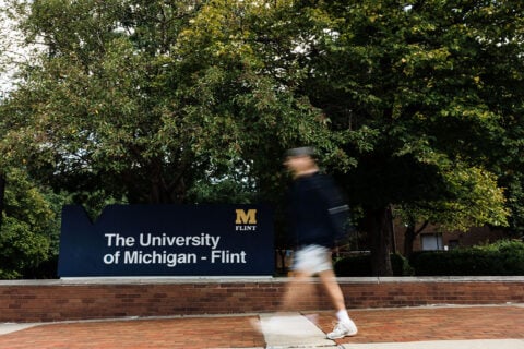 A person walks past a sign that reads "The University of Michigan - Flint," surrounded by trees.