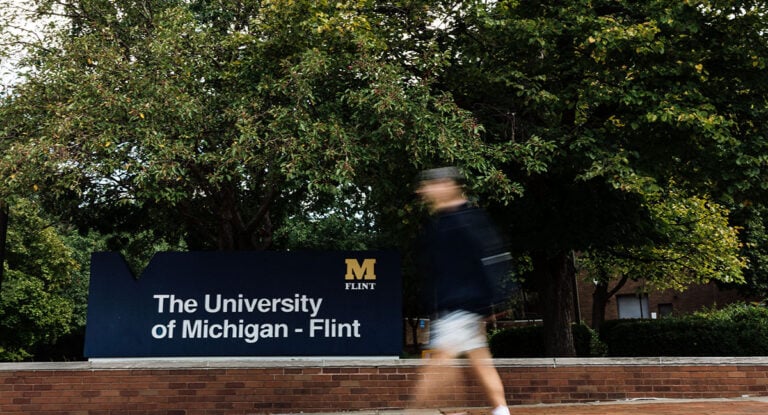 A person walks past a sign that reads "The University of Michigan - Flint," surrounded by trees.