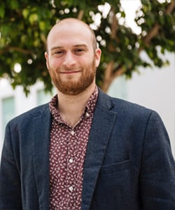 A man with a beard is smiling, wearing a dark blazer over a patterned shirt, standing outdoors with greenery in the background.