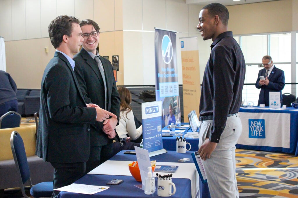 Three young men are engaged in conversation at a career fair. Two are in suits, while one is in a black shirt and light pants. Tables with promotional materials are in the background.