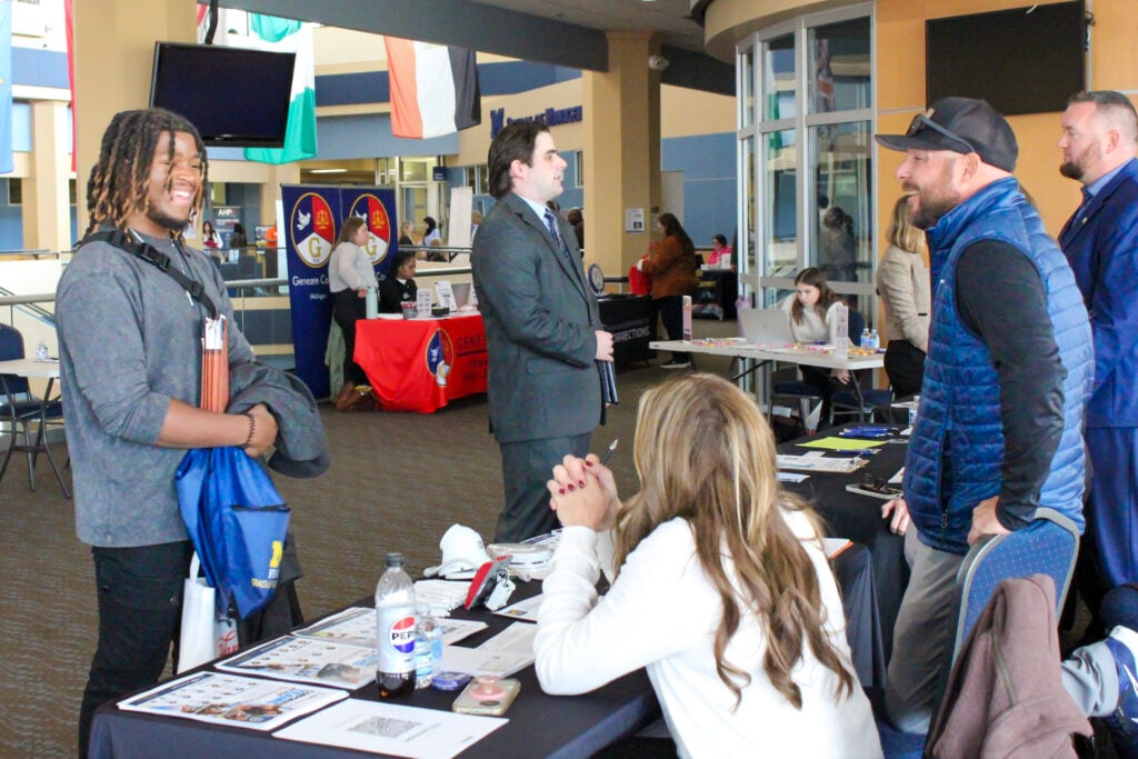 A group of people interacting at a career fair, with one young man smiling and holding materials, while others engage in conversation at tables.