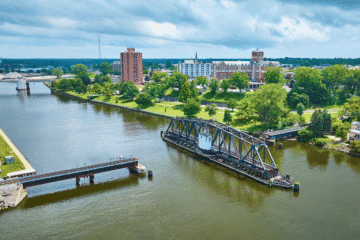 Aerial view of a river with a movable bridge, surrounded by green trees and buildings in the background under a cloudy sky.