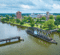 Aerial view of a river with a movable bridge, surrounded by green trees and buildings in the background under a cloudy sky.