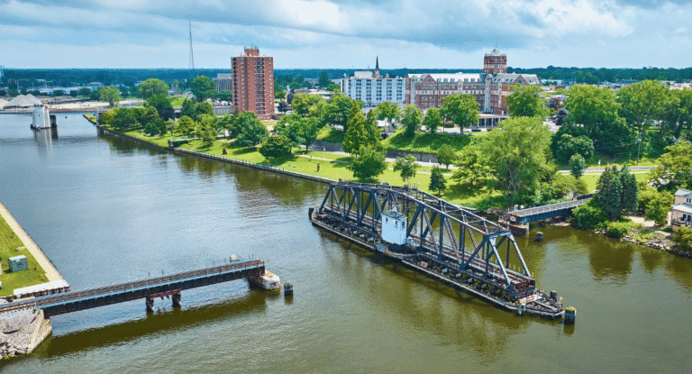Aerial view of a river with a movable bridge, surrounded by green trees and buildings in the background under a cloudy sky.