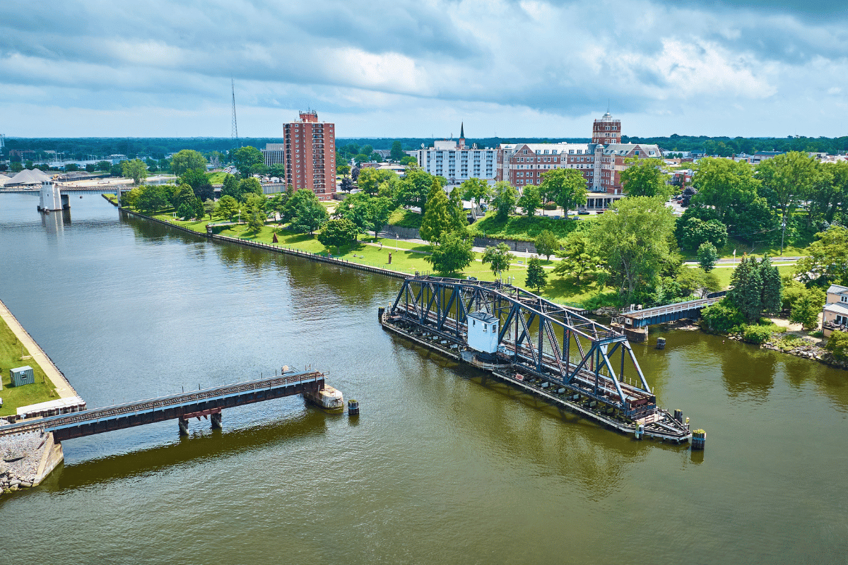 Aerial view of a river with a movable bridge, surrounded by green trees and buildings in the background under a cloudy sky.