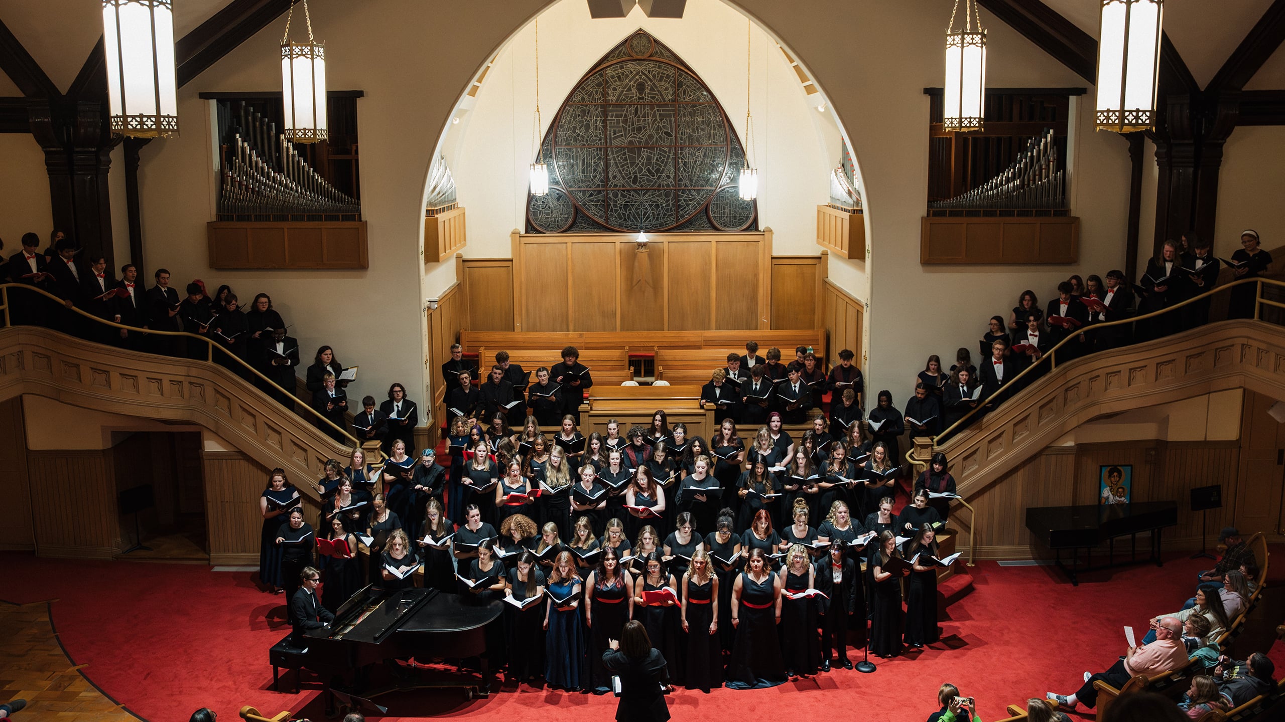 A large choir performs in a church setting, with singers in formal attire on a red carpeted stage, accompanied by a pianist. The backdrop features stained glass and an organ.