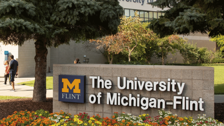 Sign for The University of Michigan-Flint, featuring a large "M" logo and surrounded by greenery and flowers.