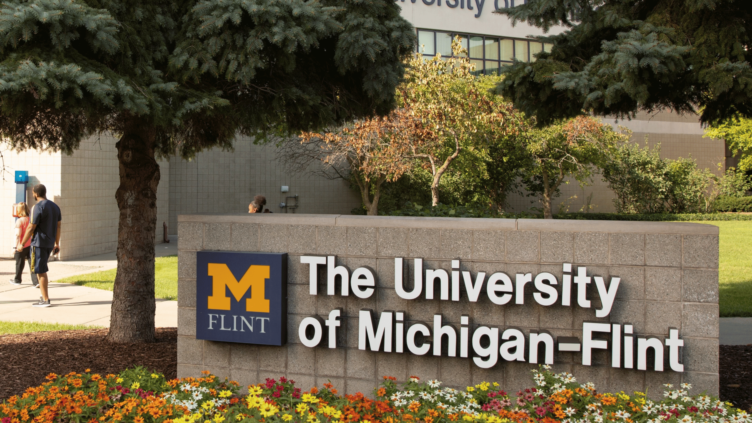 Sign for The University of Michigan-Flint, featuring a large "M" logo and surrounded by greenery and flowers.