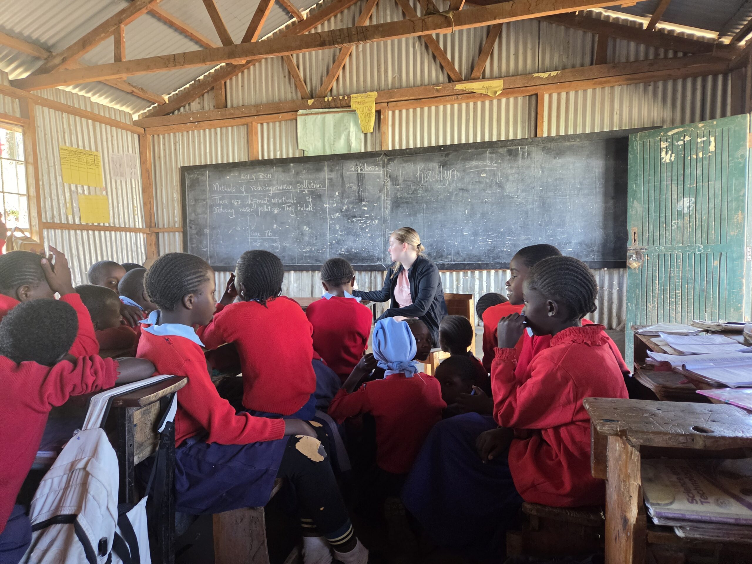 A teacher sits in front of a classroom, engaging with students wearing red uniforms. The classroom has a chalkboard and wooden desks.
