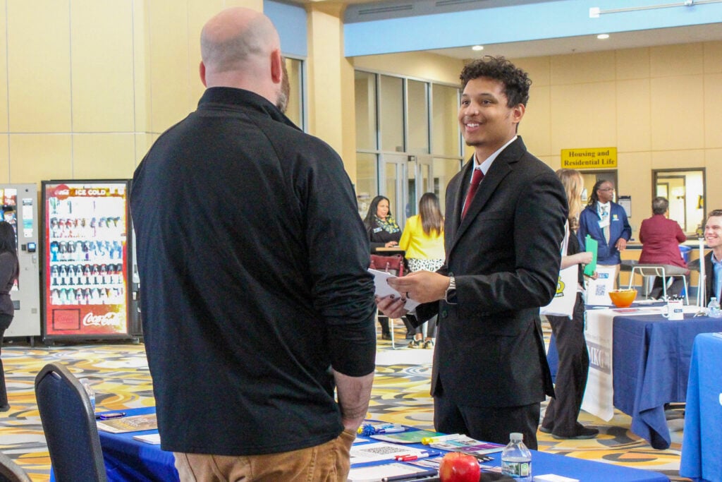 A young man in a suit smiles and holds papers while talking to another man in a black shirt at a career fair. Vending machines and tables with informational materials are in the background.