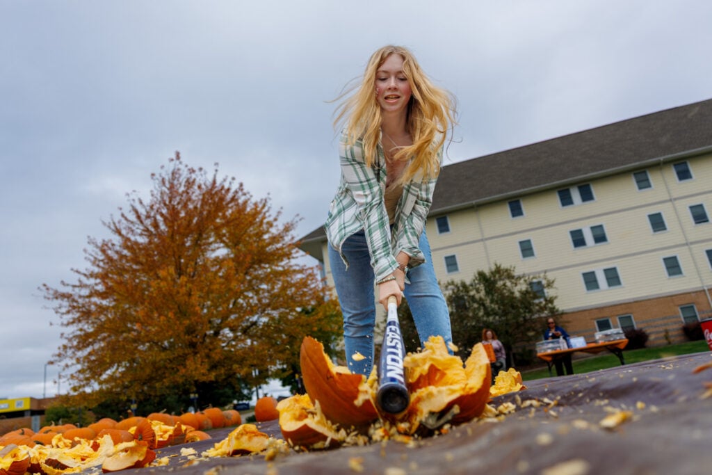 A young woman swings a baseball bat to smash a pumpkin, with pumpkin pieces scattered around her. In the background, there are trees and a building.