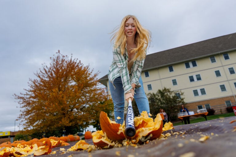 A young woman swings a baseball bat to smash a pumpkin, with pumpkin pieces scattered around her. In the background, there are trees and a building.