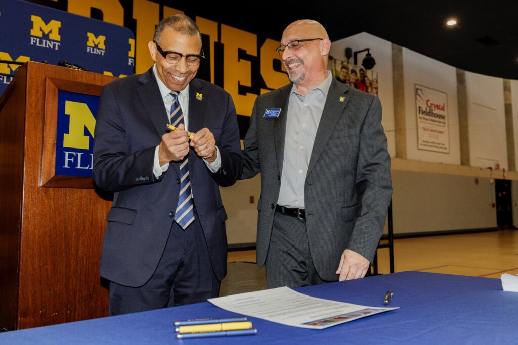 Two men are smiling and interacting at a table with a blue tablecloth. One man is holding a pen, and they appear to be engaged in a discussion. A podium with the University of Michigan Flint logo is in the background.