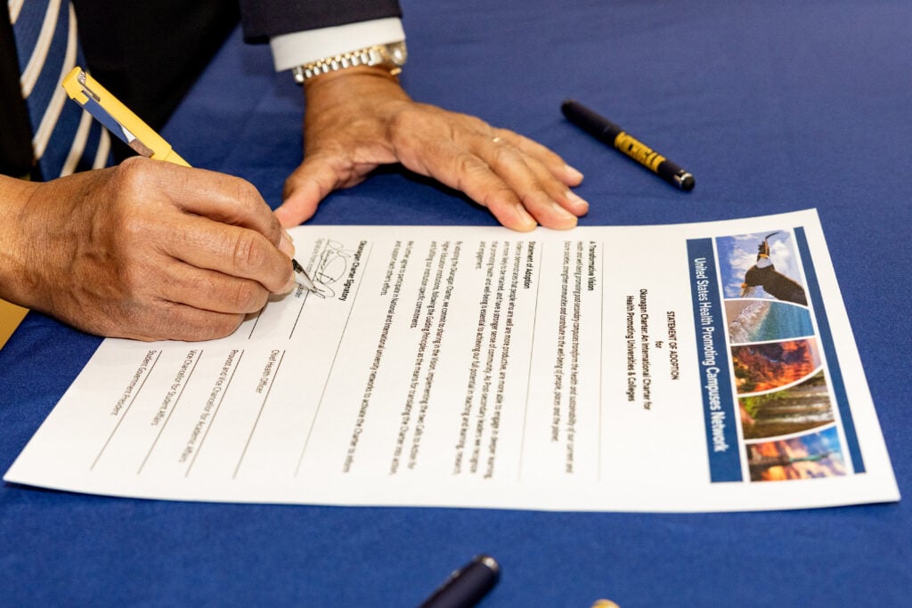 A hand is signing a document on a blue tablecloth, with a yellow pen. The document is titled "United States Health Promoting Campuses Network."