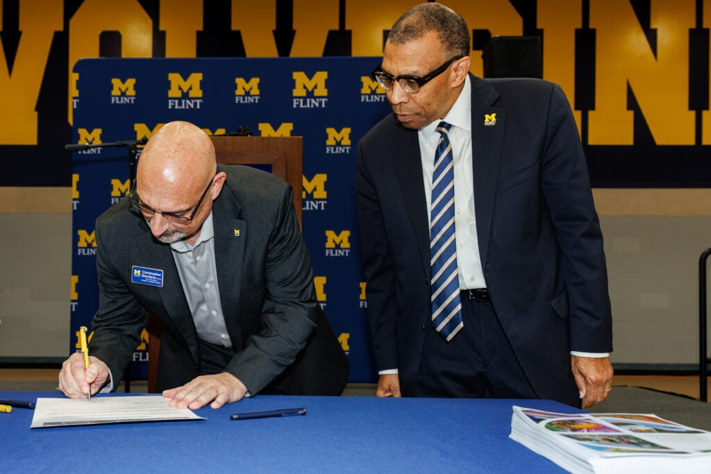 A man with glasses is signing a document at a table, while another man in a suit observes him. The background features a blue and yellow banner with "M FLINT."