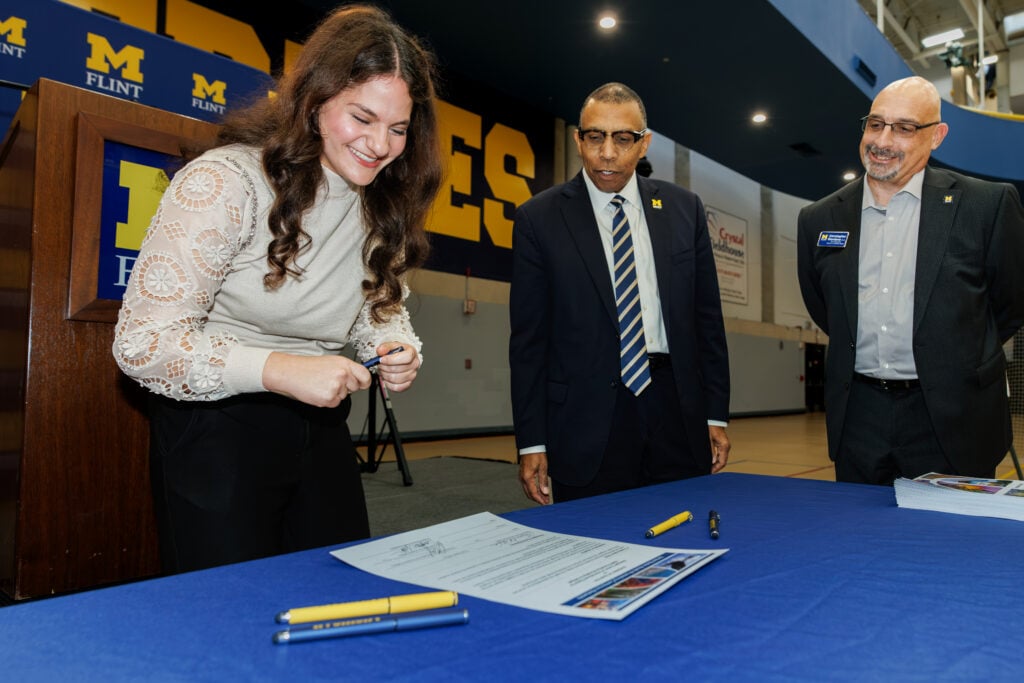 A young woman is smiling as she signs a document at a table, while two men observe her. The setting appears to be a university event.