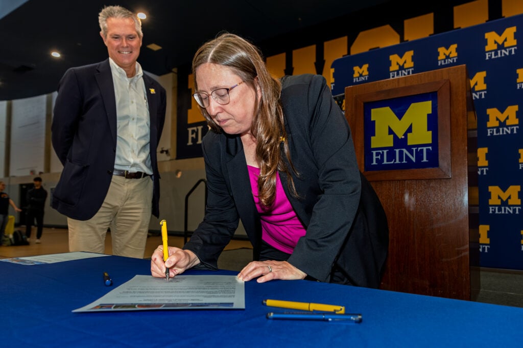 A woman in a black blazer and pink top is signing a document at a table, while a man in a suit stands smiling in the background. A podium with the "M FLINT" logo is visible behind them.