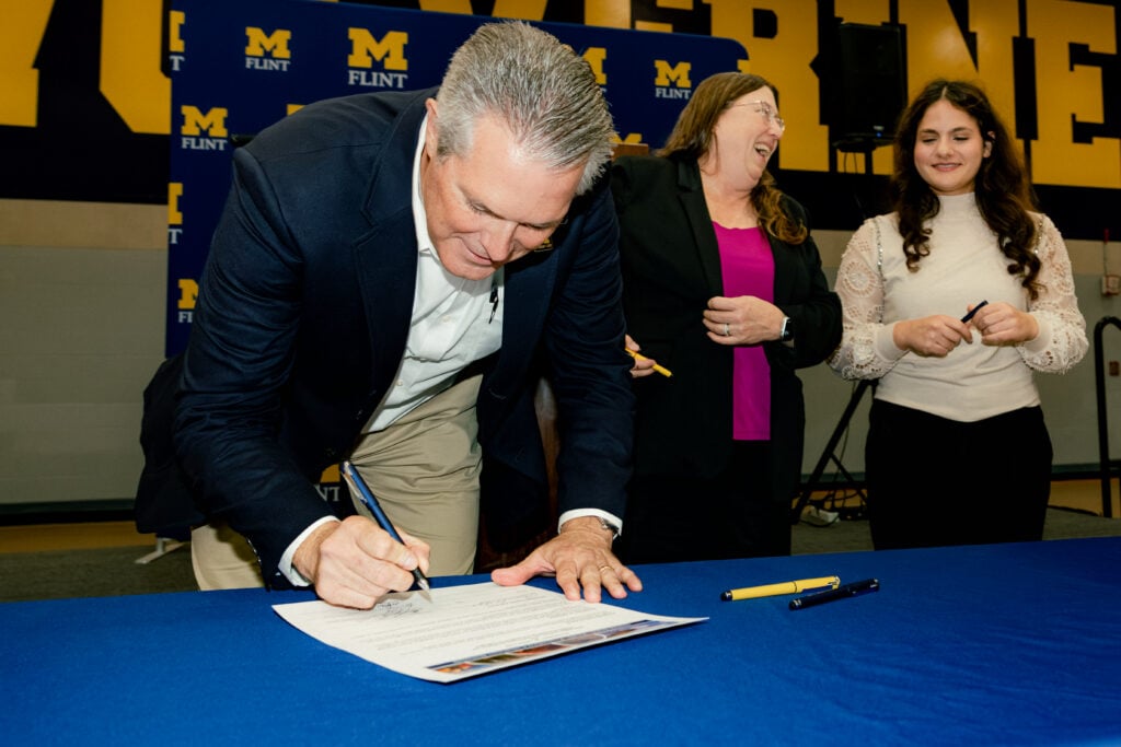 A man in a suit signs a document at a table, while two women stand nearby, smiling and watching. The background features a blue and yellow banner with "M FLINT."