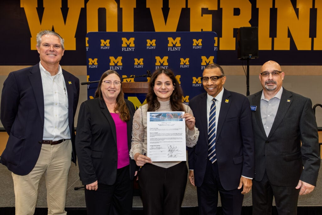 A group of five people stands together, smiling. The central figure, a young woman, holds a certificate. They are in front of a backdrop with "Wolverines" and "M FLINT" in yellow and blue.