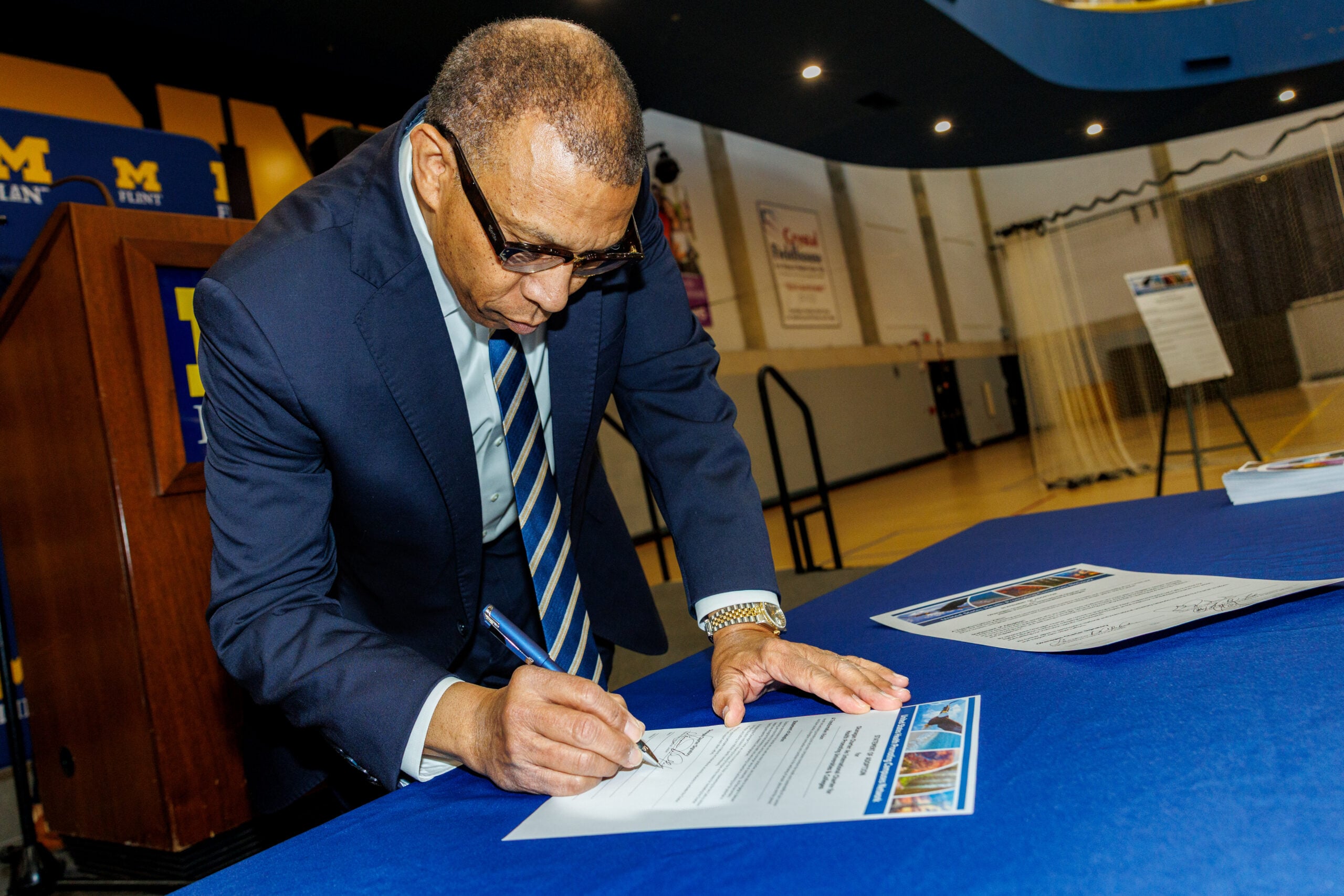 A man in a suit is signing a document on a blue table in a gymnasium, with a podium and banners in the background.